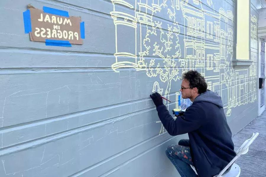 一位艺术家在教会区一栋建筑的一侧画壁画, 大楼上贴着一个牌子，上面写着“正在进行壁画”. 贝博体彩app，加利福尼亚.