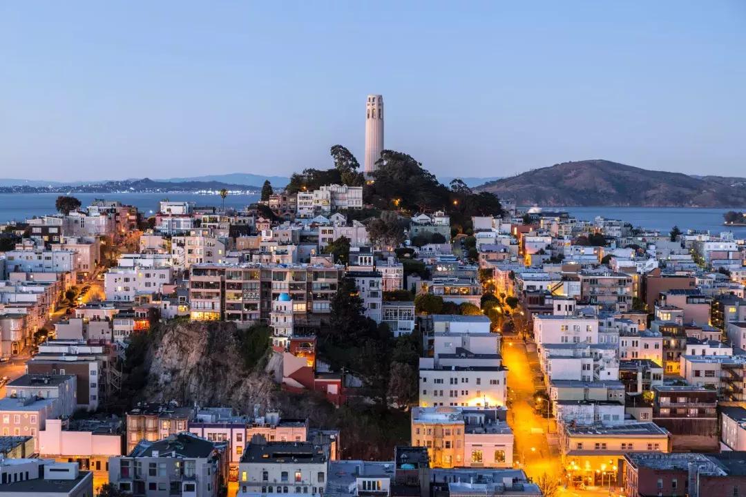 Der Coit Tower von San Francisco in der Abenddämmerung, mit beleuchteten Straßen davor und der Bucht von San Francisco dahinter.