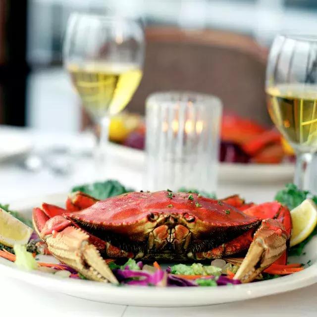 A Dungeness crab sits on a plate in a restaurant with two glasses of white wine in the background.