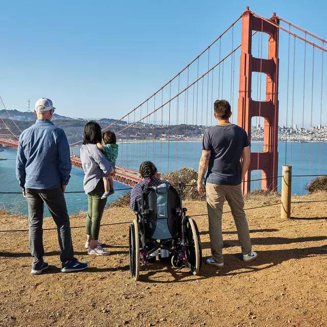 Um grupo de pessoas, incluindo uma pessoa em cadeira de rodas, é visto por trás enquanto olham para a Ponte Golden Gate de Marin Headlands.